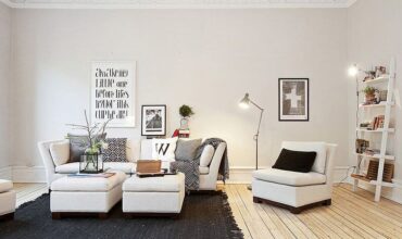 Apartamento pequeno decorado em tons de branco com sofá, poltrona, estante, tapete e quadros.