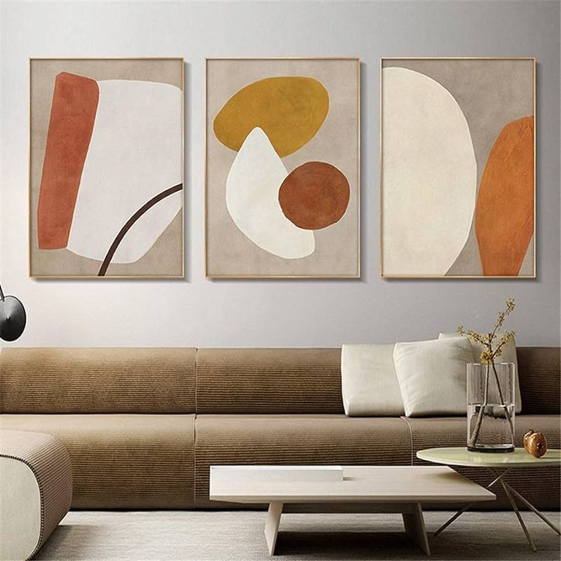 Sala de estar com sofá marrom claro e três quadros com detalhes na cor creme e laranja 